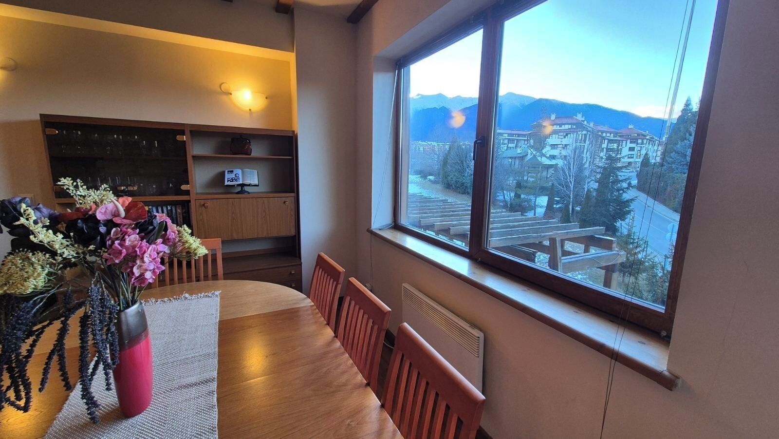 A dining area with a wooden table and chairs, a vase with flowers, and a view of a town through a large window.