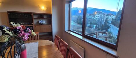 A dining area with a wooden table and chairs, a vase with flowers, and a view of a town through a large window.