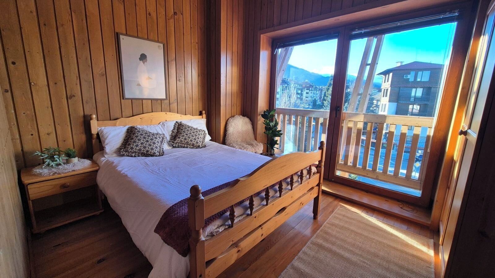 Un dormitorio de madera con cama, mesita de noche, ventana con vistas a edificios y balcón.