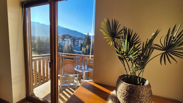 A balcony with a wooden table and chairs, a potted plant, and a view of a mountain and buildings. - 3 bedroom Penthouse only 700 meters from Ski Lift (Bansko)