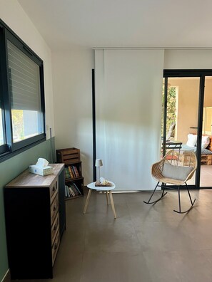 A room with a large window, a wooden desk with a tissue box, a small table with a lamp, a wicker rocking chair, and a sliding glass door leading to an outdoor area. - 5 minutes from the beach, beautiful studio in a quiet, secure residence (Grosseto-Prugna)
