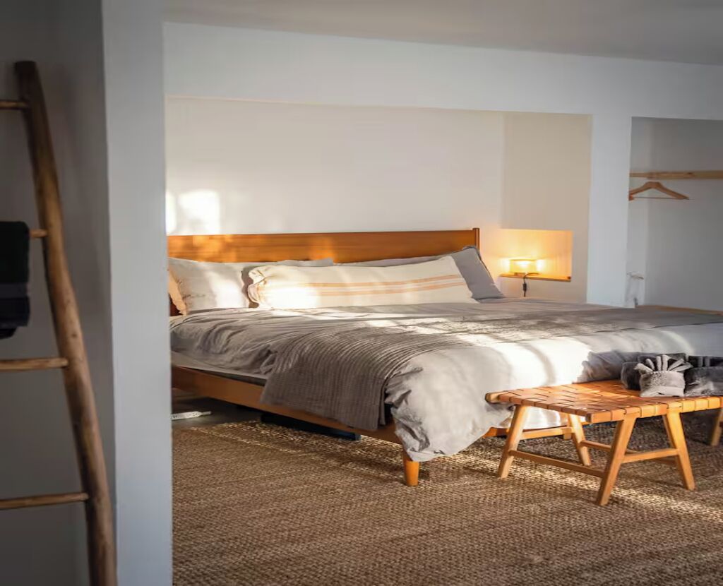 Un lit bien fait, avec une tête de lit en bois, une lampe de chevet et une petite table en bois sur laquelle est posé un chat.