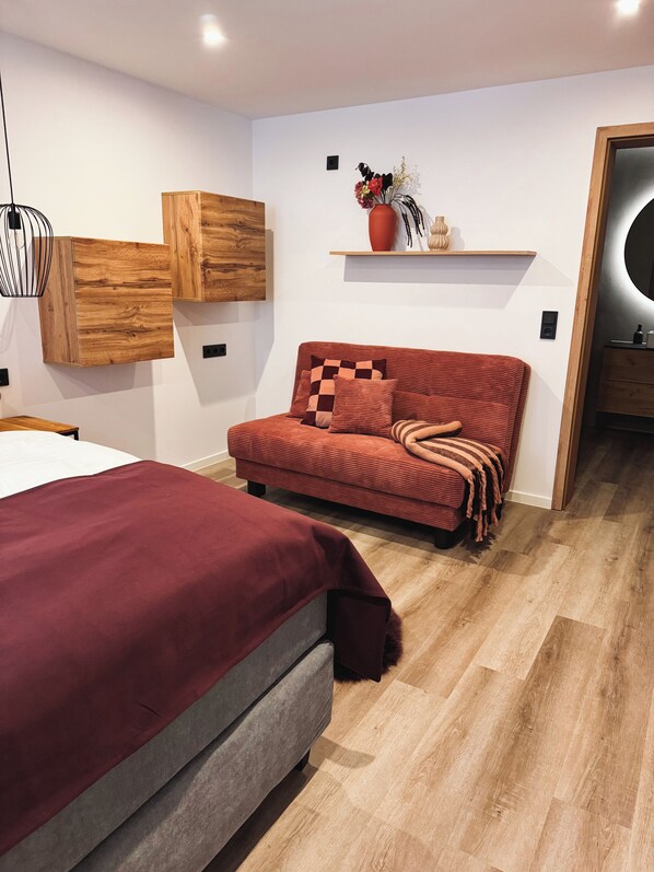 Quarto moderno com cama, sofá vermelho, painéis de parede de madeira e uma pequena estante com objetos decorativos.