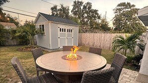 A round outdoor patio table with wicker chairs, a fire pit in the center, and a small shed in the background. - Dunedin Palm Retreat •1 Mile to Downtown Dunedin •Walk to Pinellas Trail & Beach (Clearwater)