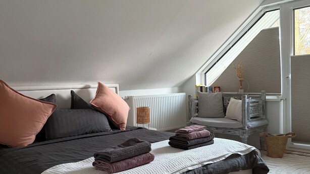 Un dormitorio con techo inclinado, cama con ropa de cama oscura, mesita de noche con lámpara y ventana con persianas.