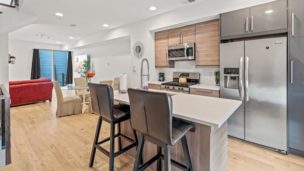 A modern kitchen with stainless steel appliances, a central island with bar stools, and a dining area with a red sofa. - Fancy 4BR W/ Hot Tub (Philadelphia)
