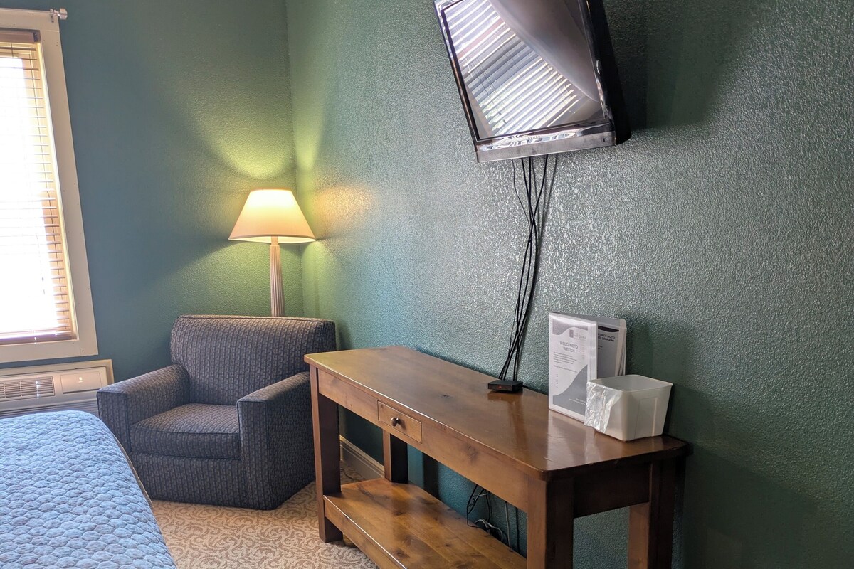 A room with a bed, a chair, a desk, a lamp, and a television mounted on the wall.