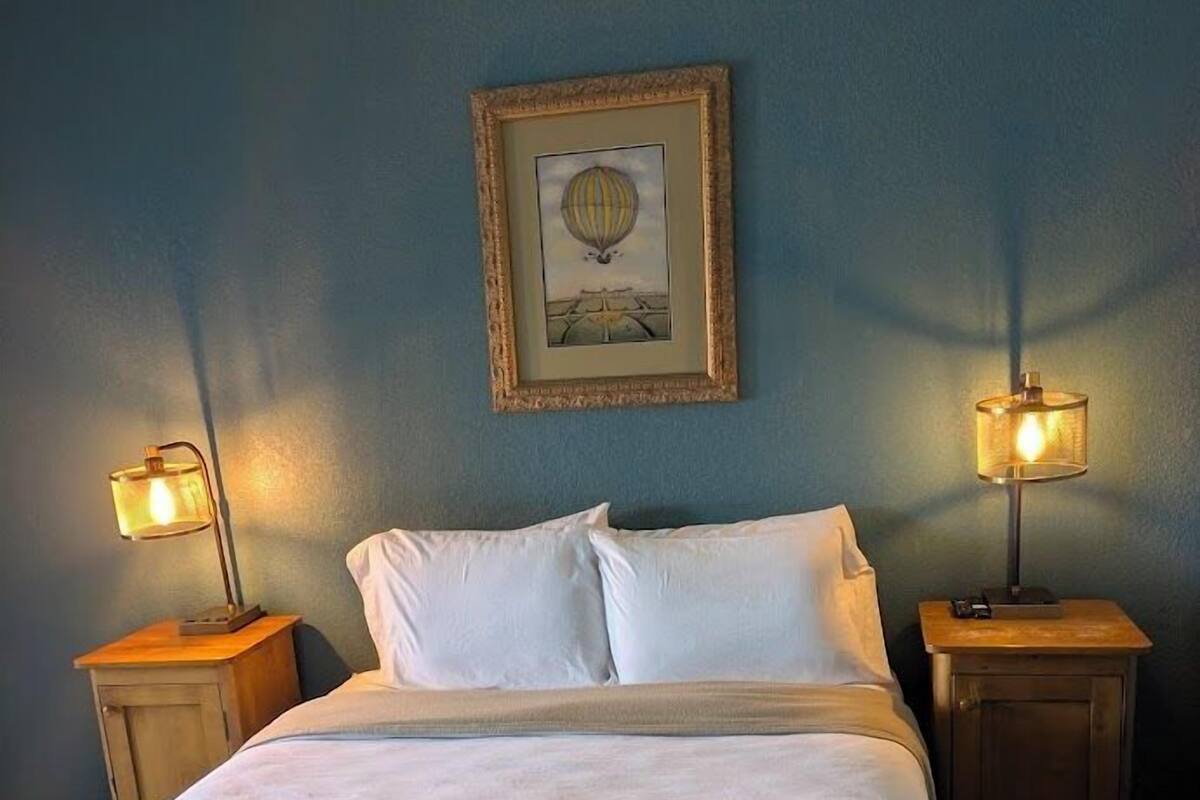 Un letto con lenzuola bianche, due comodini con lampade, un quadro appeso al muro e una parete blu.