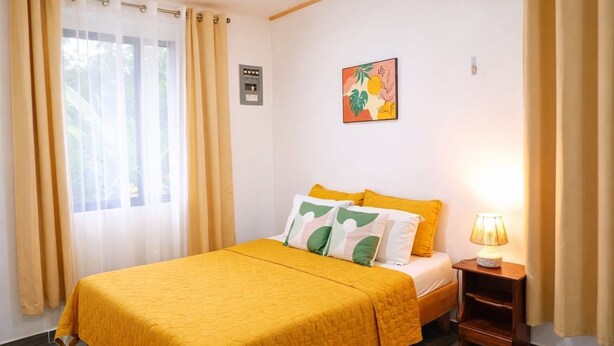Habitación de hotel con cama, cortinas, lámpara de noche, enchufe en la pared y un cuadro enmarcado.