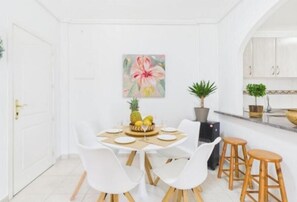 A white dining area with a round table, white chairs, and wooden stools. There is a painting on the wall and potted plants. - Casa Ellen in Los Alcazares Spanien (Los Alcázares)