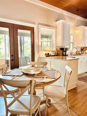 Una cocina moderna con armarios blancos, una mesa de comedor de madera con individuales y vistas al exterior a través de amplios ventanales.
