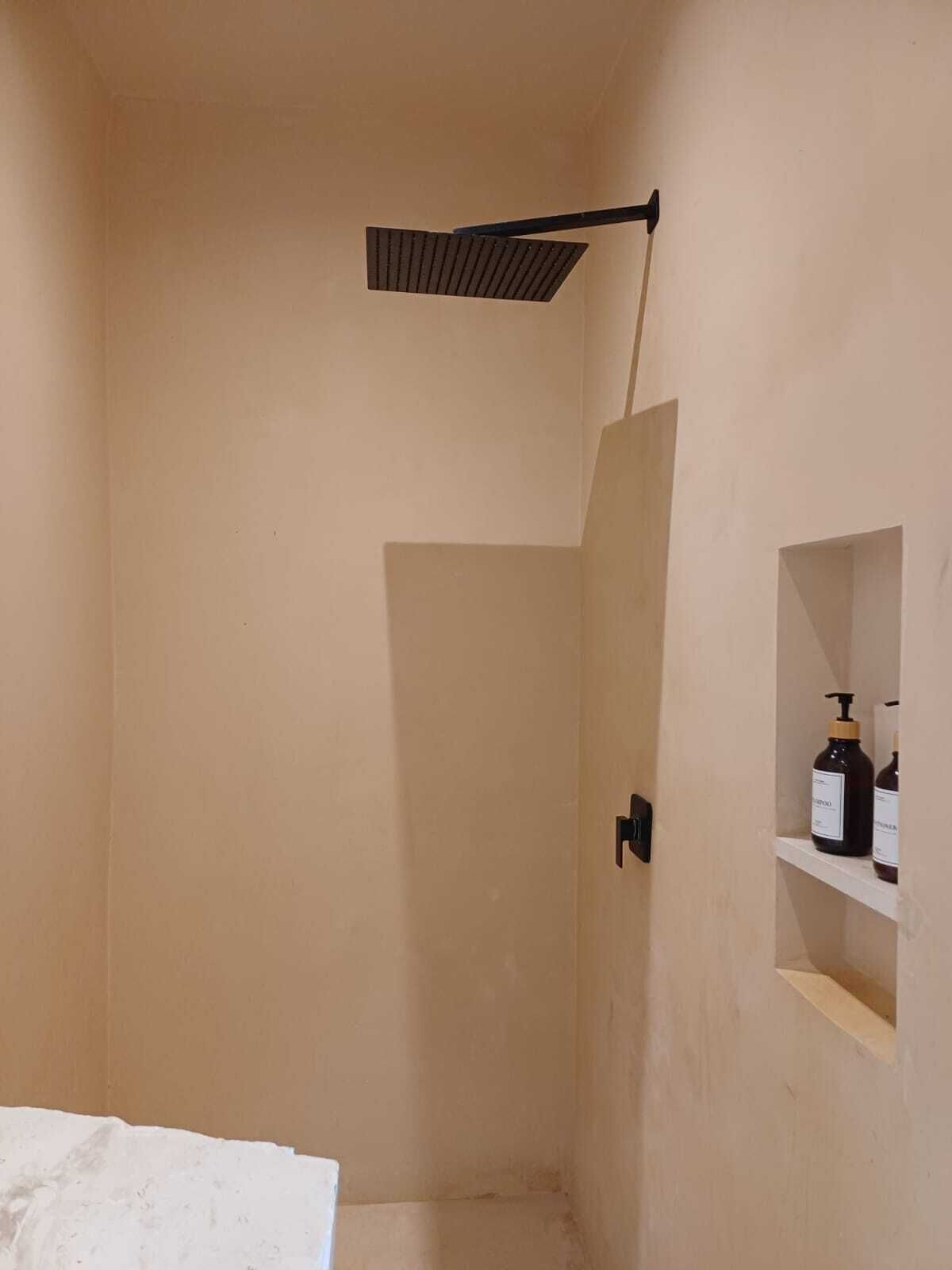 Eine Dusche mit schwarzem Griff, ein Ablagefach mit Toilettenartikeln und eine beige Wand.
