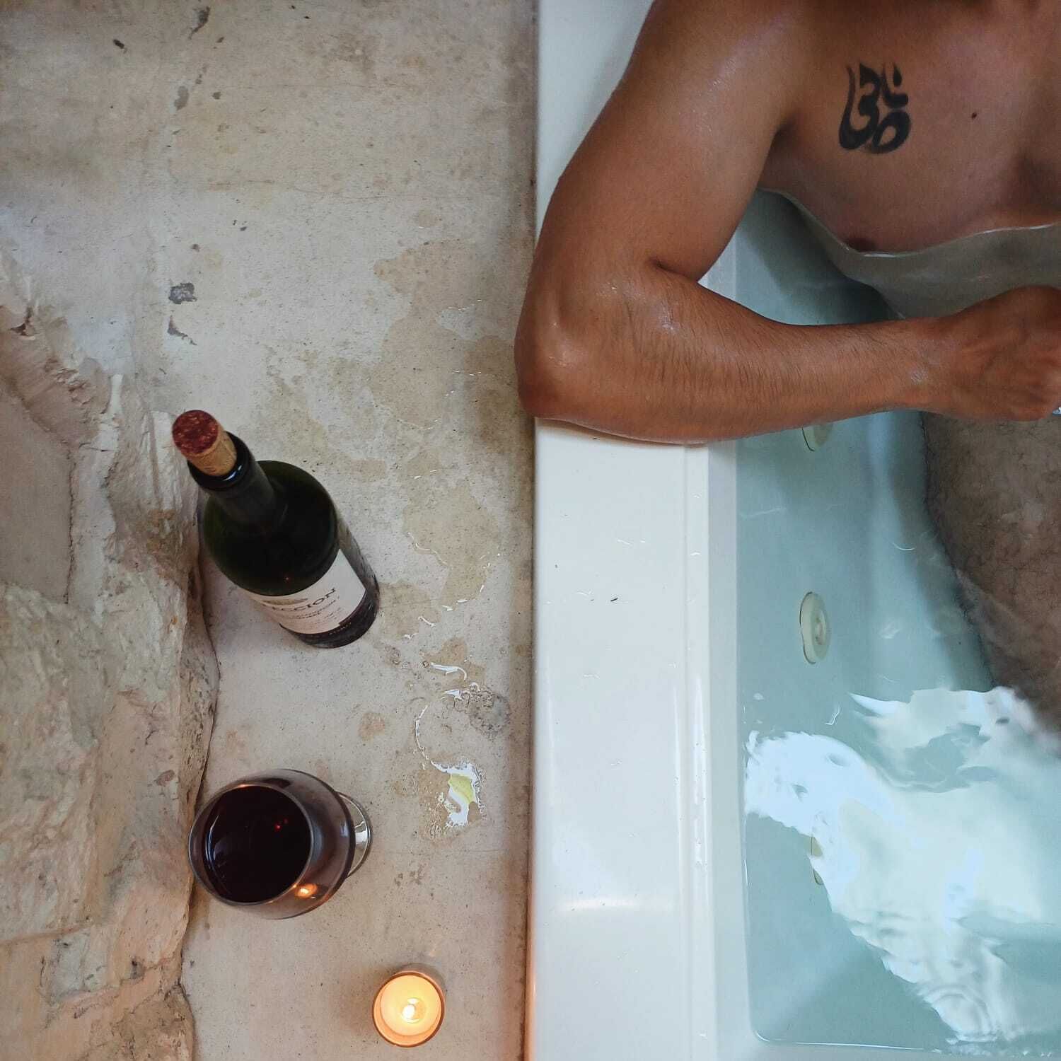 Una bañera con una persona, una botella de vino, una copa de vino y una vela encendida.