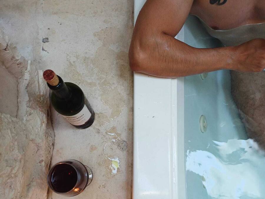 Una bañera con una persona, una botella de vino, una copa de vino y una vela encendida.