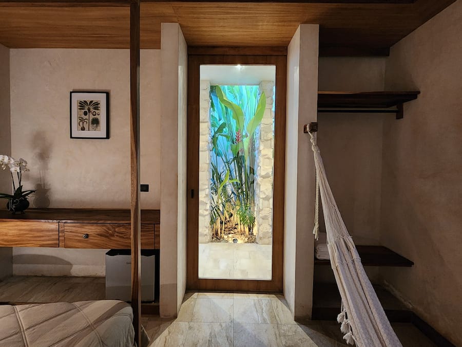 Una habitación con un escritorio de madera, una hamaca, una cama y una puerta con un panel de vidrio tropical.