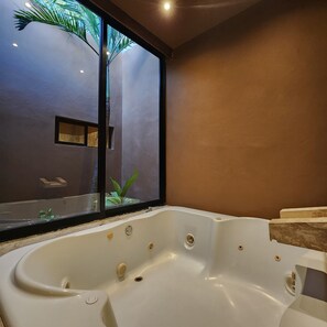 A modern bathroom with a large bathtub, a window with a view of palm trees, and a small shelf with toiletries.