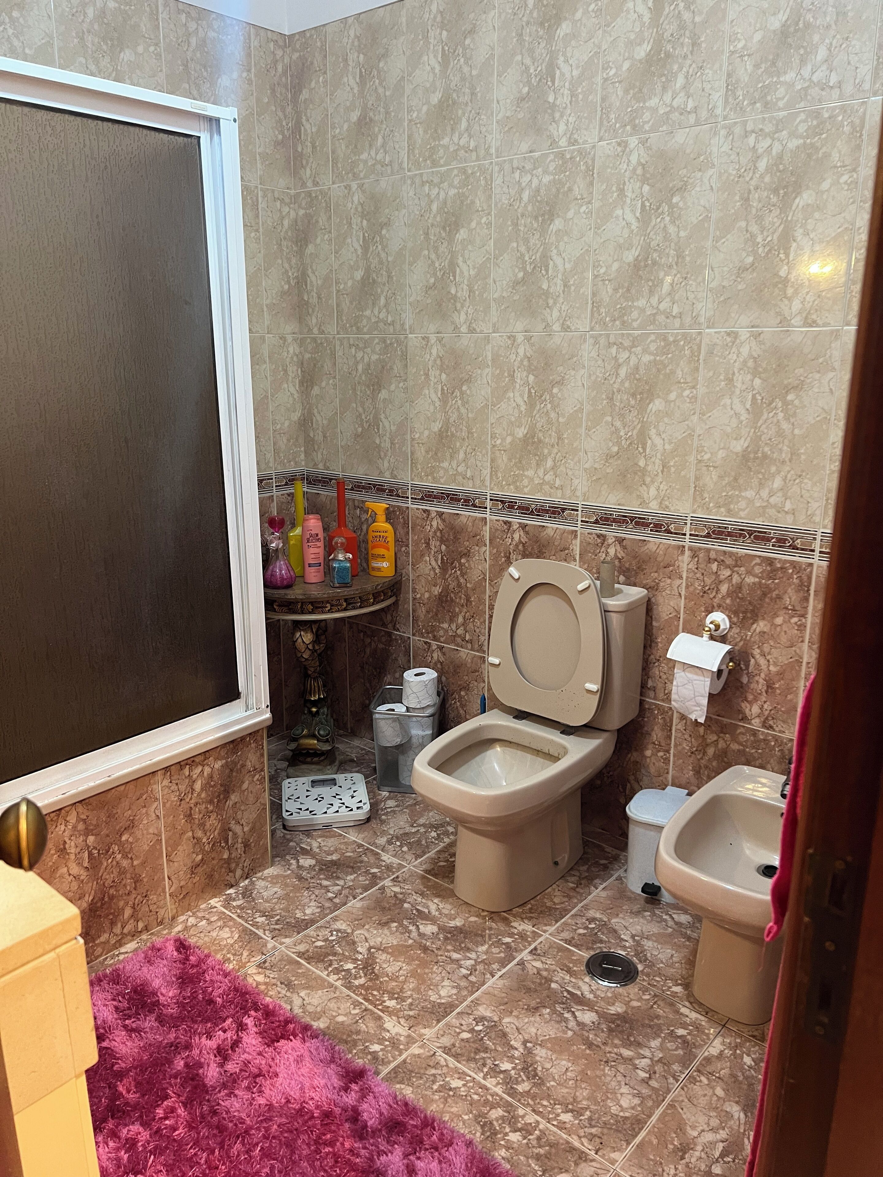A bathroom with a toilet, bidet, and sink, marble tiles, and a purple rug.