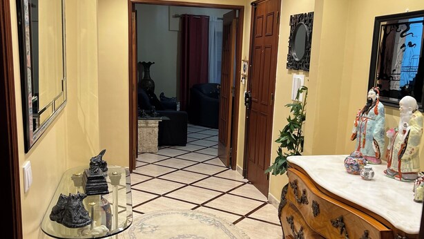 Un couloir doté d’un sol carrelé, une table en verre servant de présentoir pour des sculptures, une commode en bois avec un miroir, et une porte donnant sur une autre pièce.