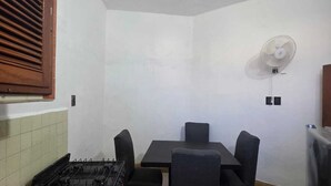 A compact kitchen with a gas stove, a dining table with four chairs, a wall-mounted fan, and a wooden window shutter. - 2 bedroom apartment near parque dela plancha park merida yucatan (Mérida)