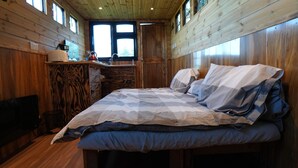 Interior de una cabaña de madera con una cama, una mesita y un ventanal.