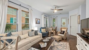 A living room with a beige sofa, a coffee table, and a television on a wooden cabinet. There is a ceiling fan, a dining table with chairs, and a window with blinds. - Waterfront Community with Private Pier + Pool (Rockport)