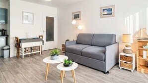 Ett modernt vardagsrum med en grå soffa, ett matbord med stolar, ett litet vitt bord med krukväxter och ett köksområde i bakgrunden.