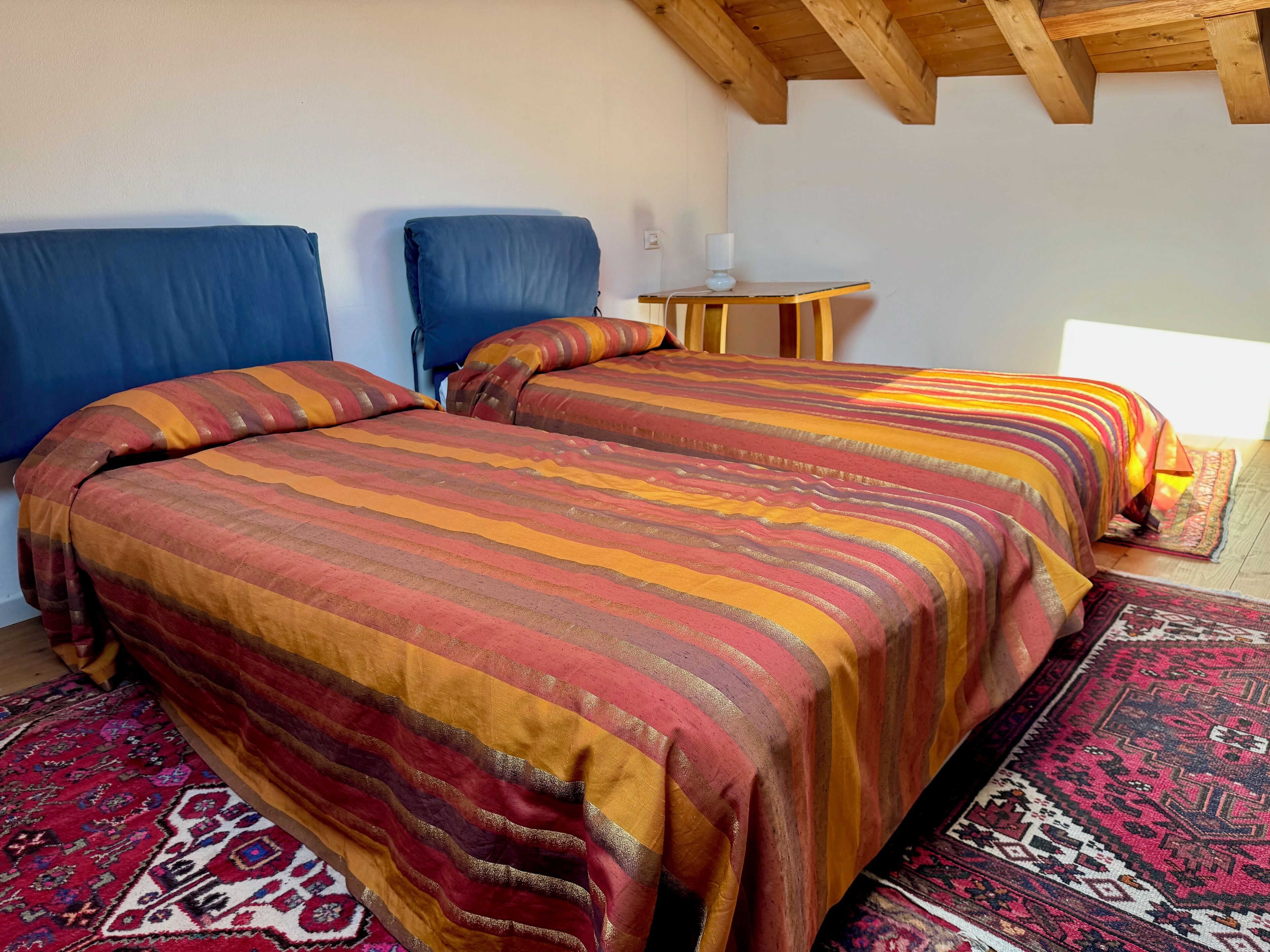 A room with two beds, a small table with a lamp, and a patterned rug.