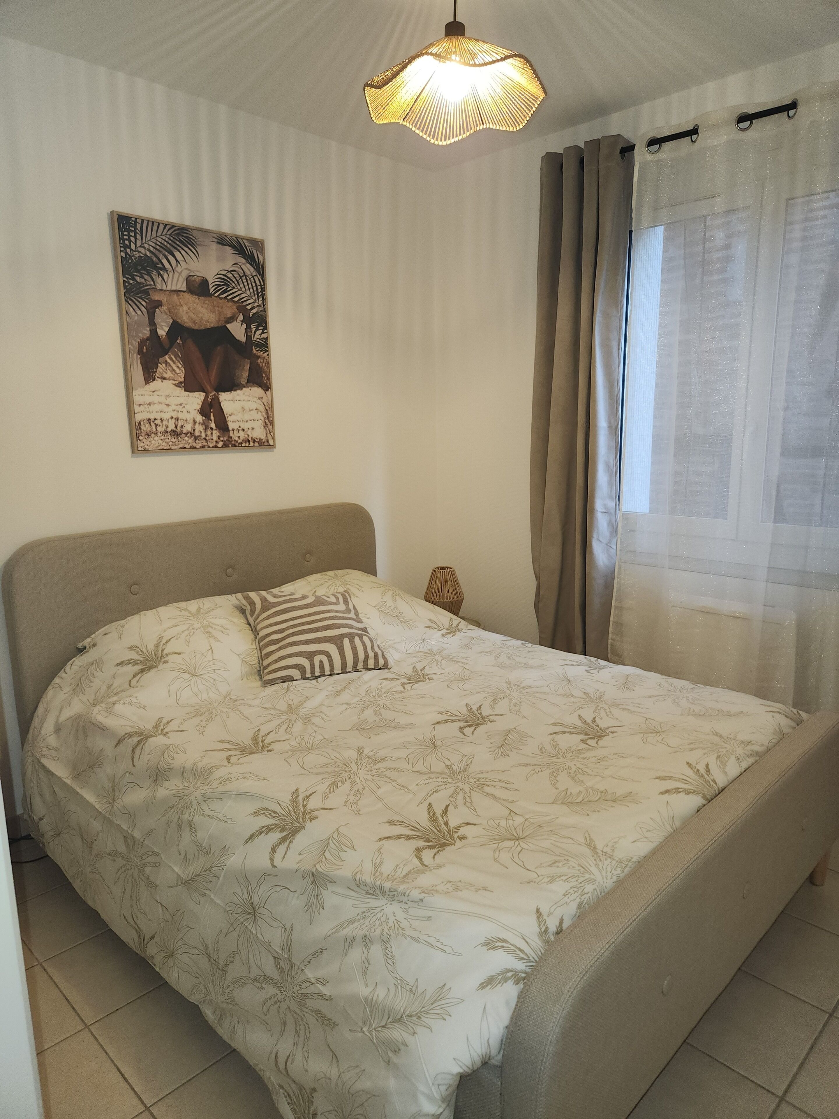 Ein Schlafzimmer mit einem Bett, einer Pendelleuchte, einem Wandbild und einem Fenster mit Vorhängen.