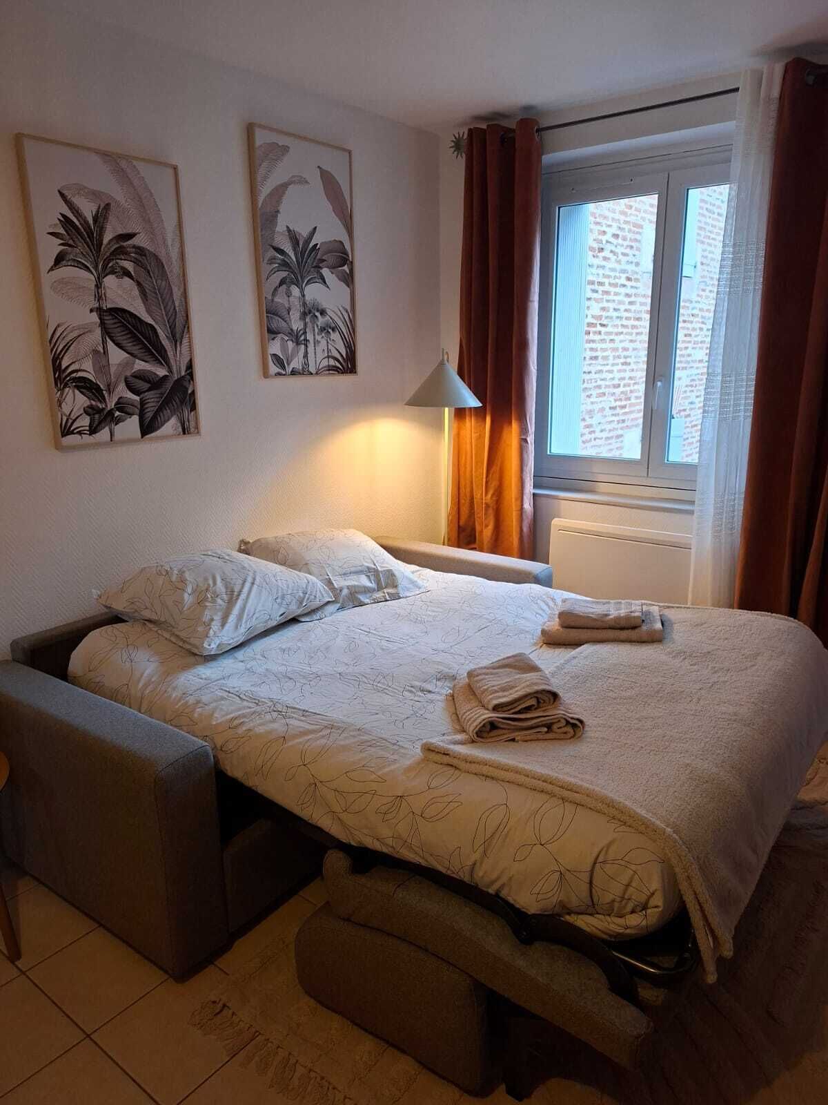 A hotel room with a bed, a bedside table, a lamp, a window with curtains, and two wall-mounted artworks.