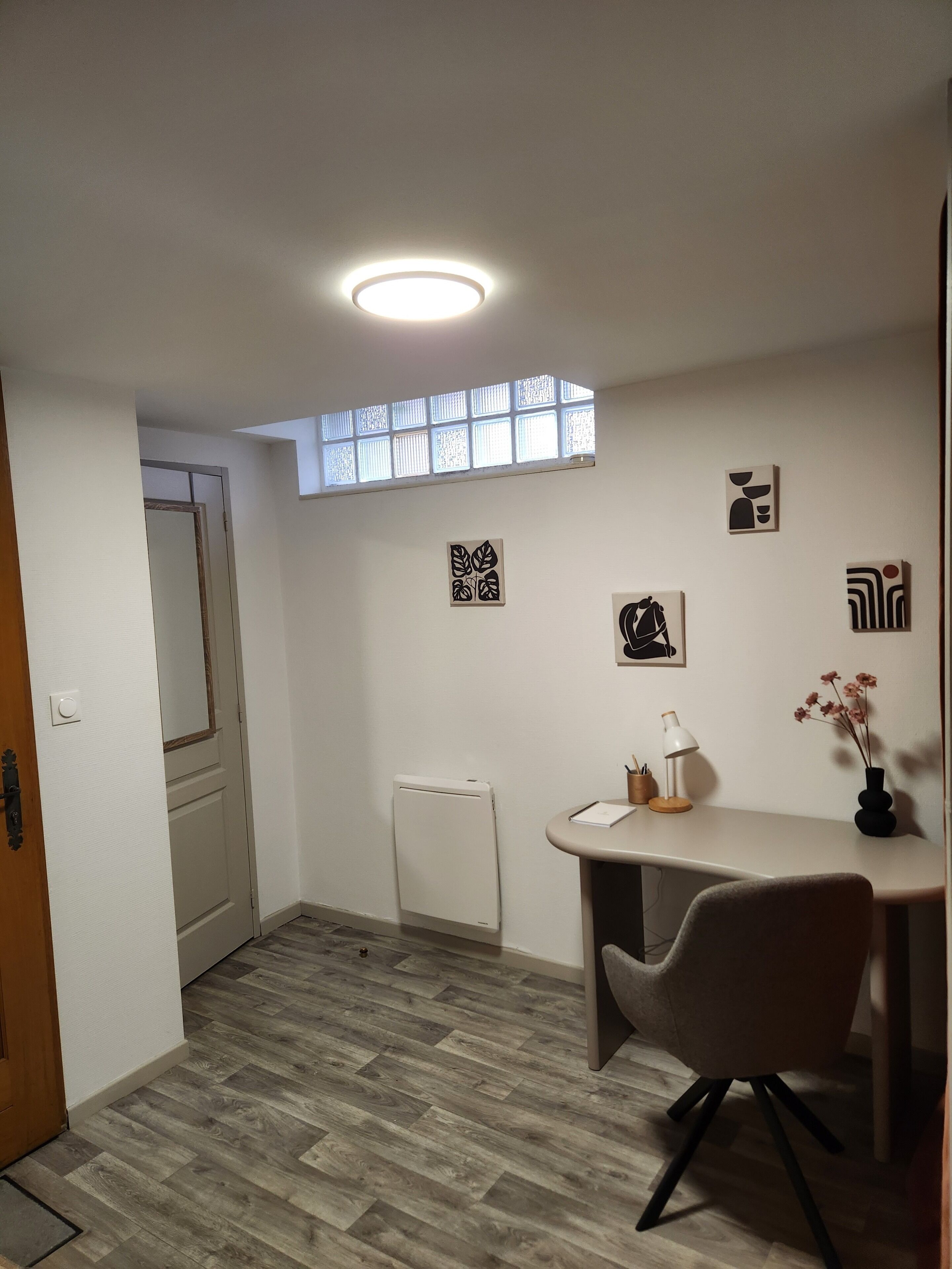 A modern room with a wooden floor, a white door, a radiator, a desk with a chair, and artwork on the walls.