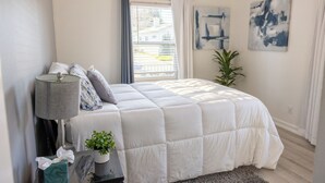 A bedroom with a bed, a bedside table with a lamp, a potted plant, and artwork on the walls.