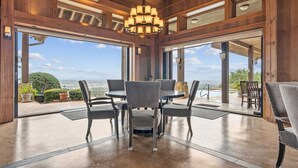 Un vaste espace dédié à la salle à manger, avec une grande table et des chaises, de grandes fenêtres offrant une vue sur le paysage, et un lustre suspendu au plafond.