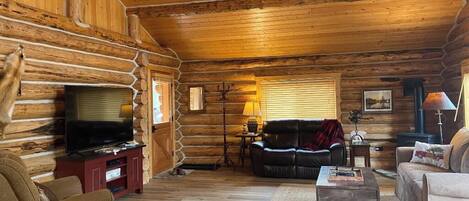 A log cabin interior with a wooden floor, a television on a stand, a sofa, a coffee table, and a fireplace.