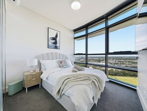 A modern bedroom with a large window, a bed with white bedding, a nightstand, and a wall-mounted picture. - The Sky Perch- Walking distance to Accor stadium and Qudos bank Arena. (Sydney Olympic Park)