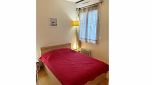 A bedroom with a wooden bed, a red blanket, a bedside table, a lamp, and a window with curtains. - 2-room apartment Maisons Laffitte/Paris (Maisons-Laffitte)