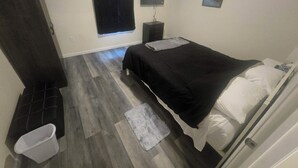 A bedroom with a bed, a nightstand, a wall-mounted TV, a dark wardrobe, and a small white trash bin. - A warm, inviting home tucked into one of Michigan’s most scenic border towns. (Sault Ste. Marie)