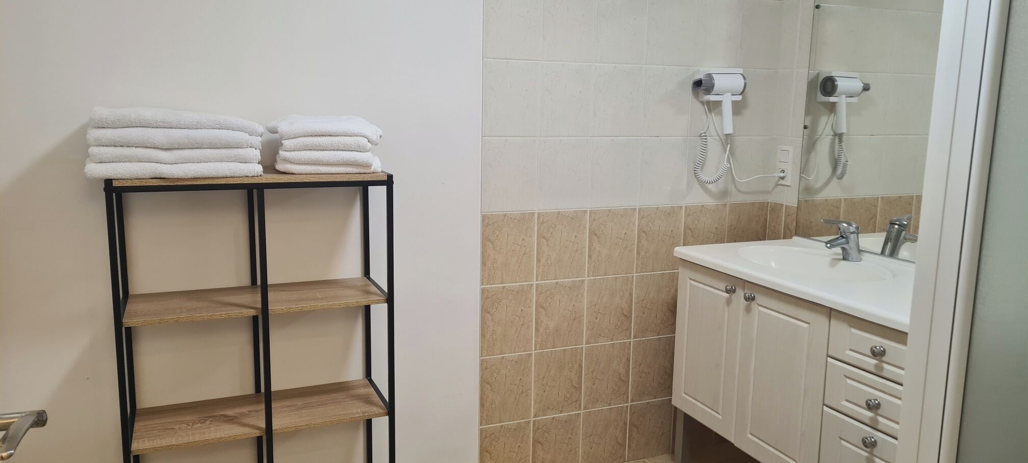 Un baño con un lavamanos blanco, un espejo y un secador de pelo montado en la pared. Hay un estante con toallas dobladas y una repisa de madera debajo.