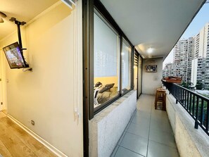 Ein Balkon mit einem an der Wand befestigten Fernseher, einem kleinen Tisch und Blick auf hohe Gebäude.