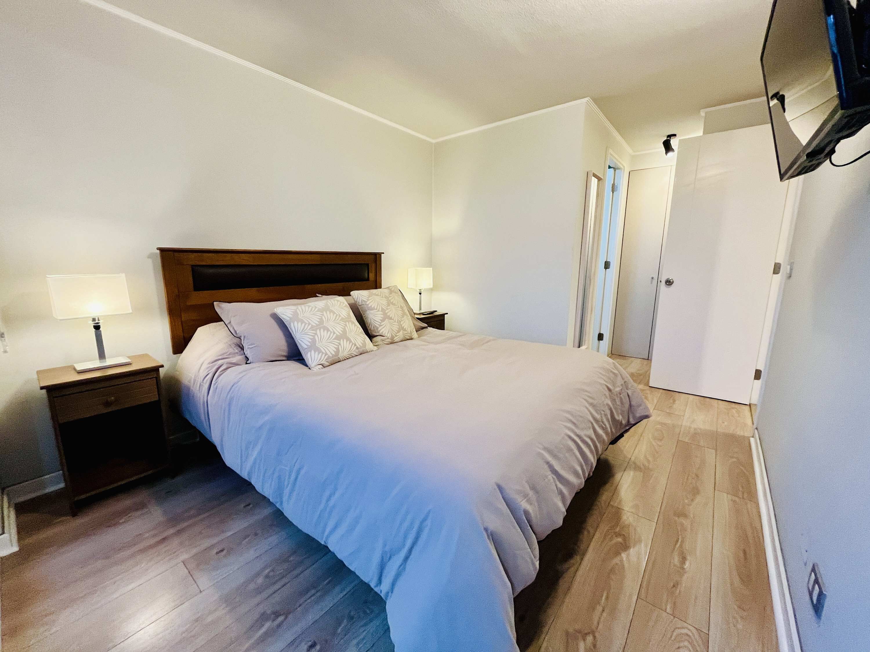 Un dormitorio con cama, mesitas de noche, un televisor montado en la pared y una puerta que da a otra habitación.