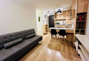 Ein kompakter Wohnraum mit grauem Sofa, einer kleinen Essecke mit zwei Stühlen und einer Küchenzeile mit Holzschränken.