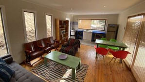 A living room with a sofa, a dining table with chairs, a kitchen area with a window, and a large window providing natural light.