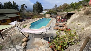 A rectangular swimming pool with a hammock and lounge chairs, surrounded by a stone border and greenery.