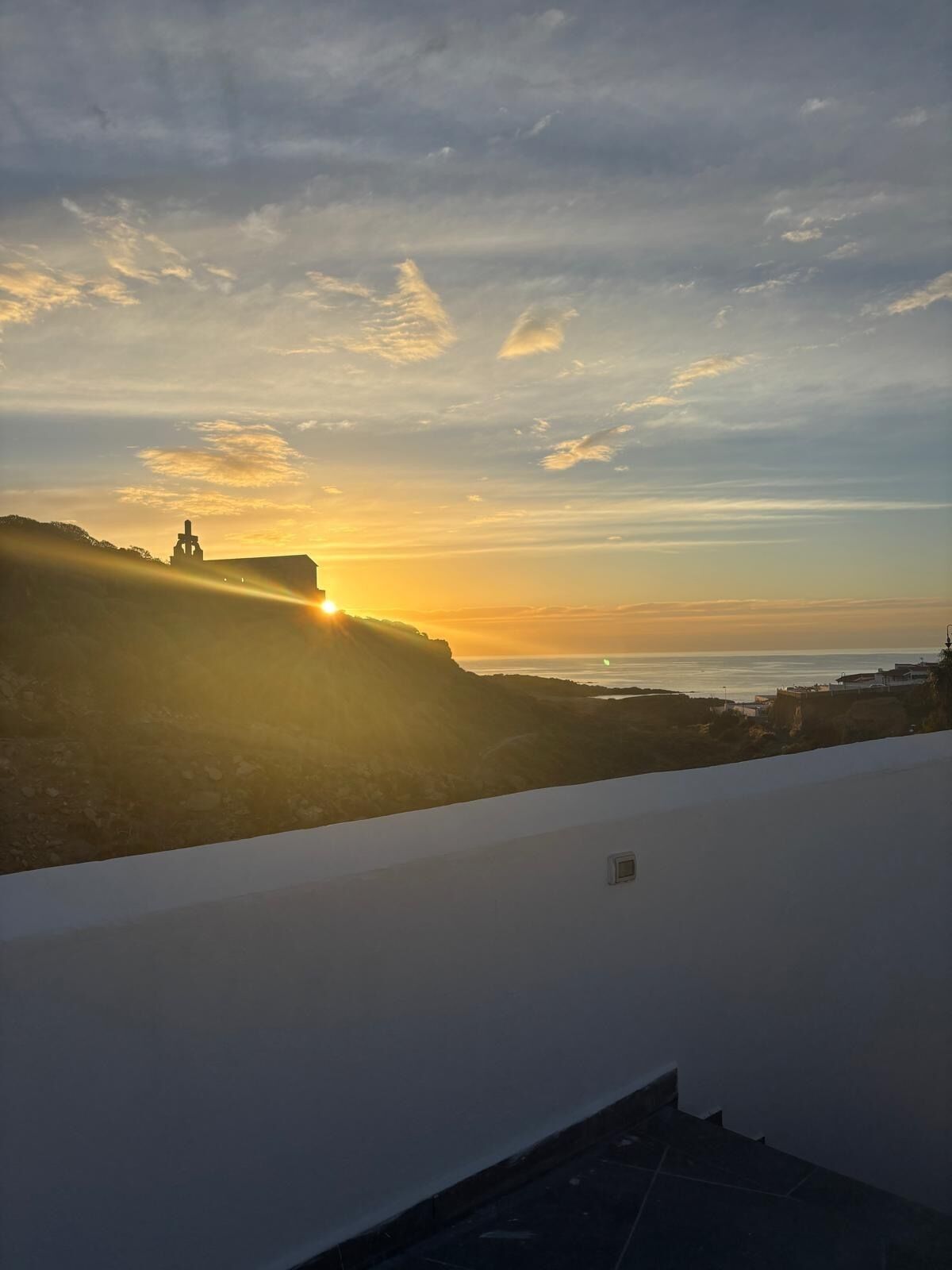 Vista do pôr do sol a partir de um terraço no telhado, com paisagem costeira, silhueta de um edifício imponente e linha costeira ao longe.