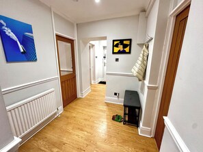 A hallway with wooden flooring, white walls, and a radiator. There are two doors, one with a wooden frame and another with a white frame. A black bench is against the wall, and a pair of shoes is on the floor.