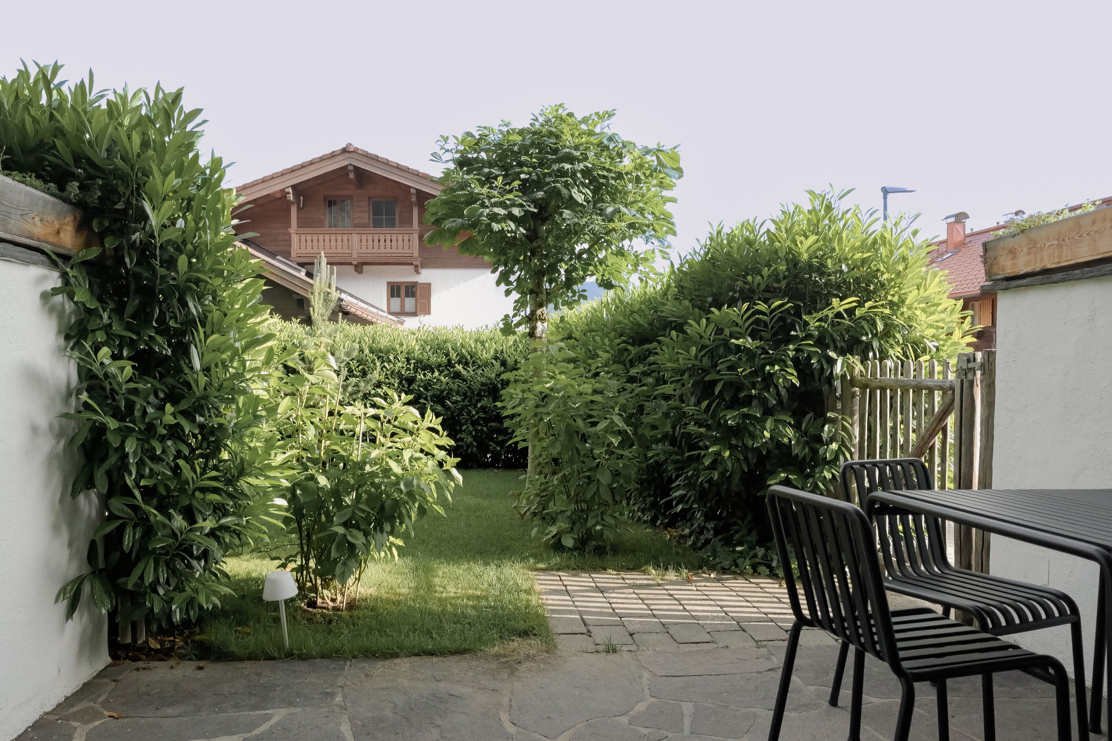 Een terras met een tafel en stoelen, omgeven door weelderige beplanting en een witte muur.