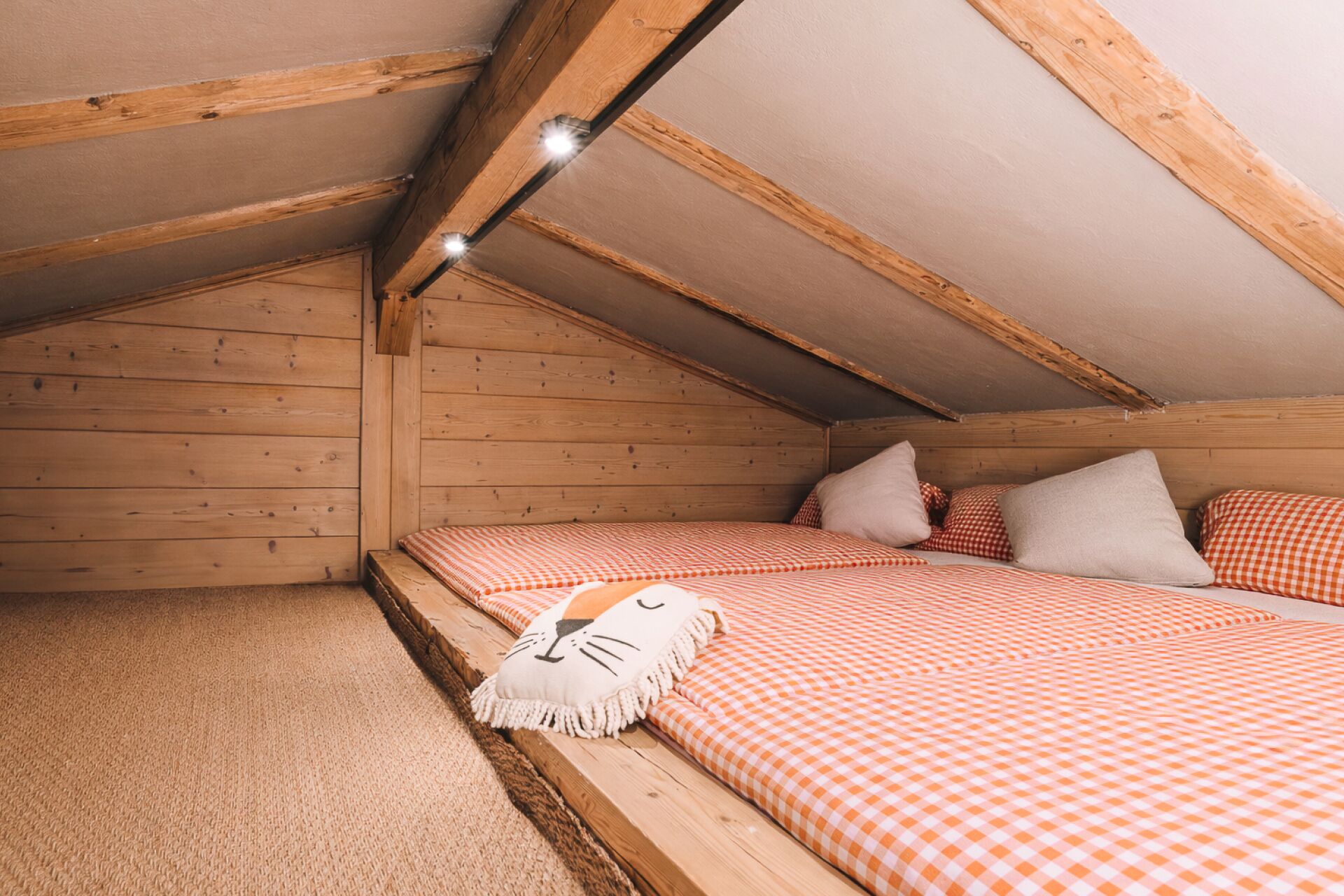 A cozy attic bedroom with wooden beams, a checkered bedspread, and a cat-themed pillow.