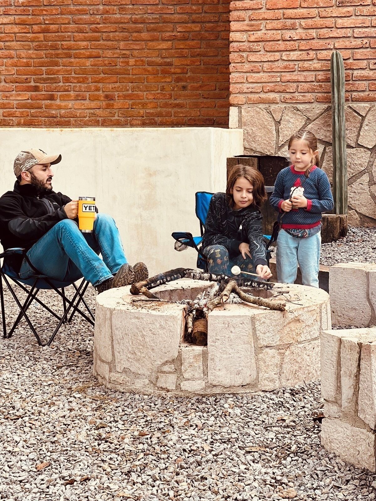 A man sitting on a folding chair, a child playing with a toy, and another child holding a toy, in front of a stone fire pit with a cactus in the background.