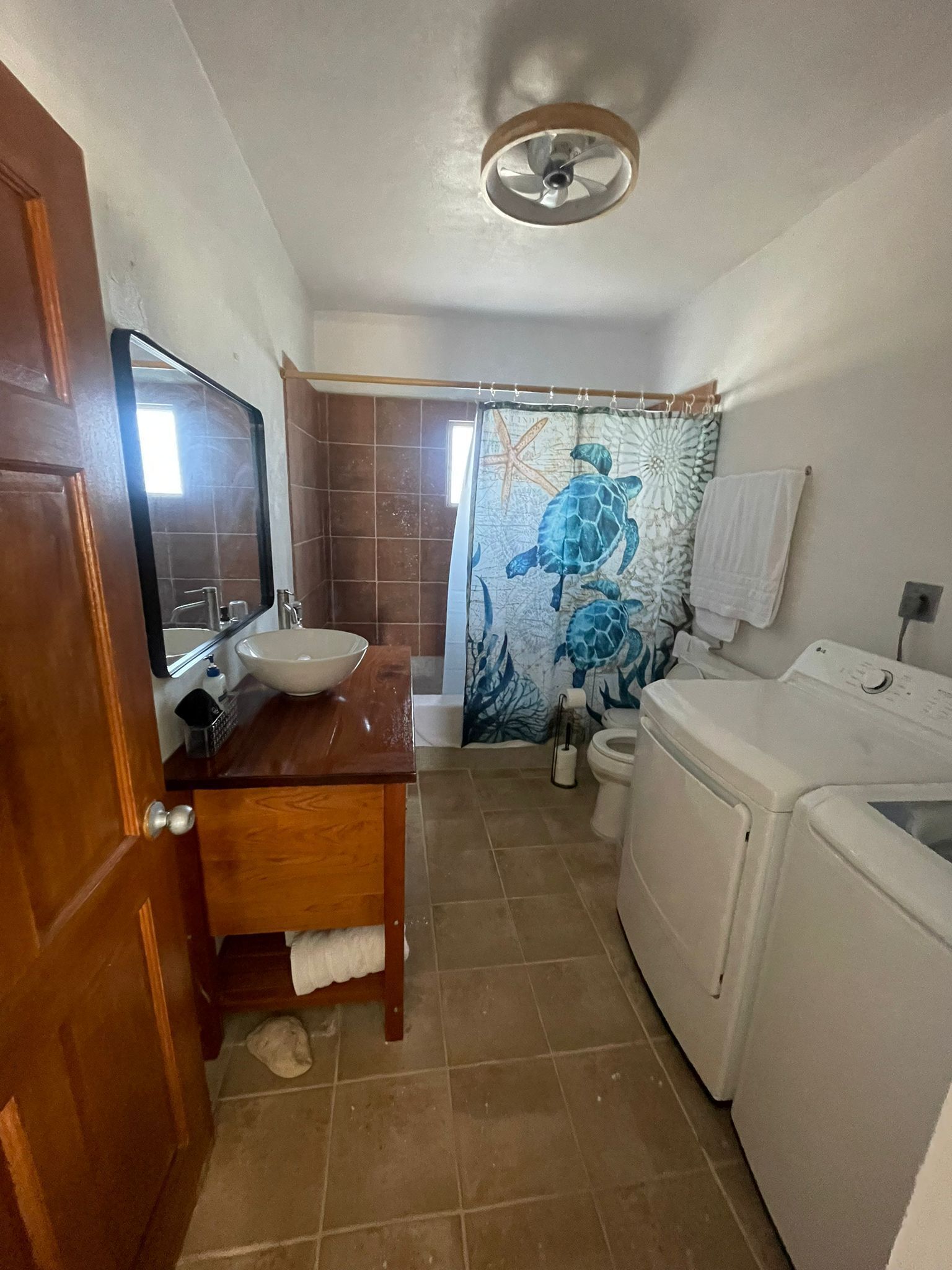 Un baño con tocador de madera, lavamanos, espejo, cortina de ducha con diseño de tortuga marina y lavadora.