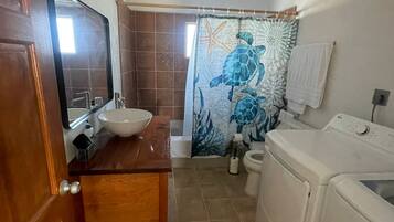 Un baño con tocador de madera, lavamanos, espejo, cortina de ducha con diseño de tortuga marina y lavadora.
