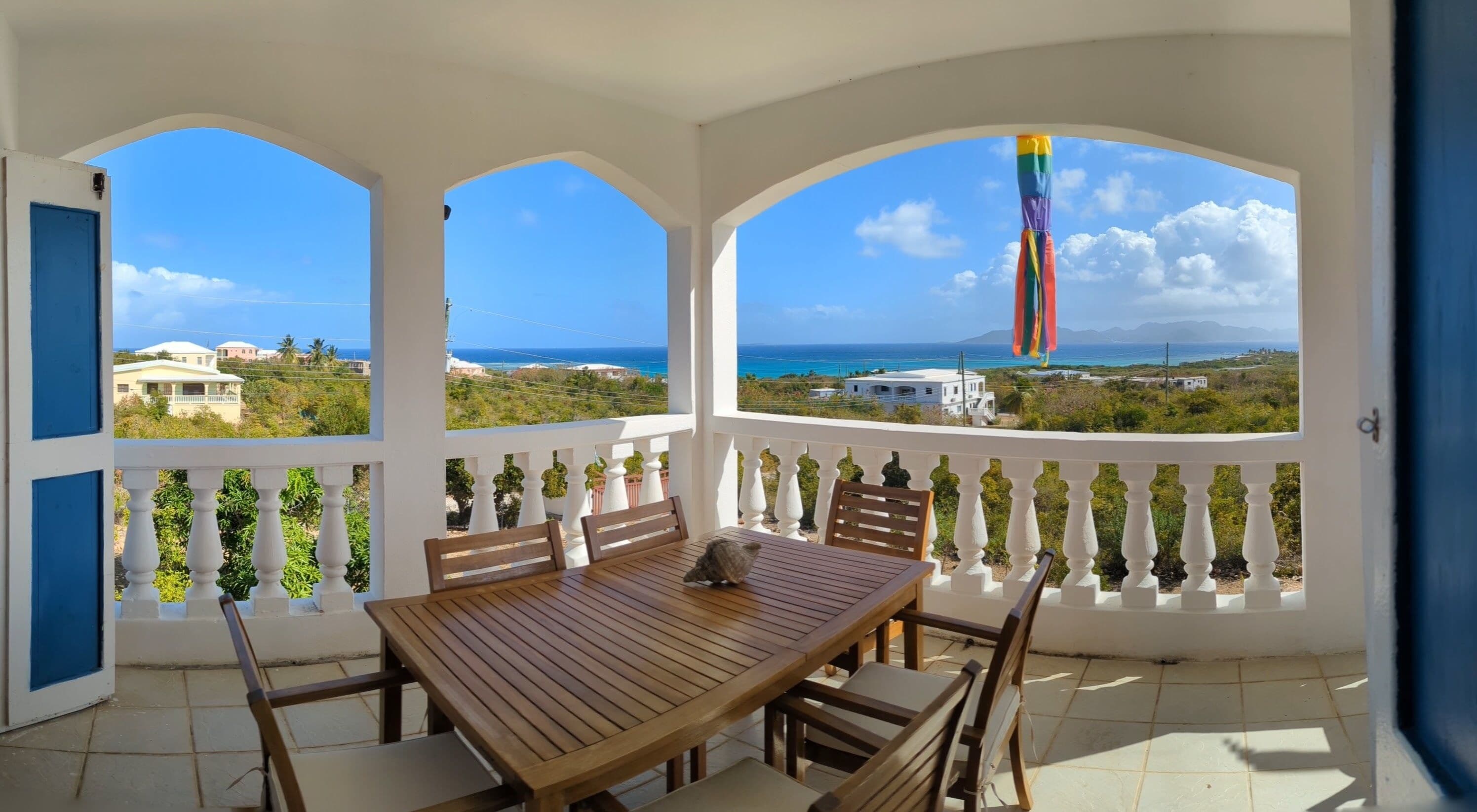 Un porche cubierto con mesa y sillas de madera, balaustradas blancas y vistas al mar y edificios.
